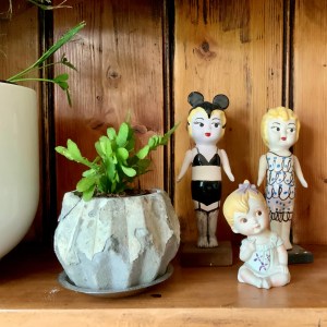 Photo of two plants and three porcelain figures: a seated girl, a standing kewpie in a dress, and another kewpie with underwear and mouse ears, to illustrate all life stages of a woman and all issues covered in therapy